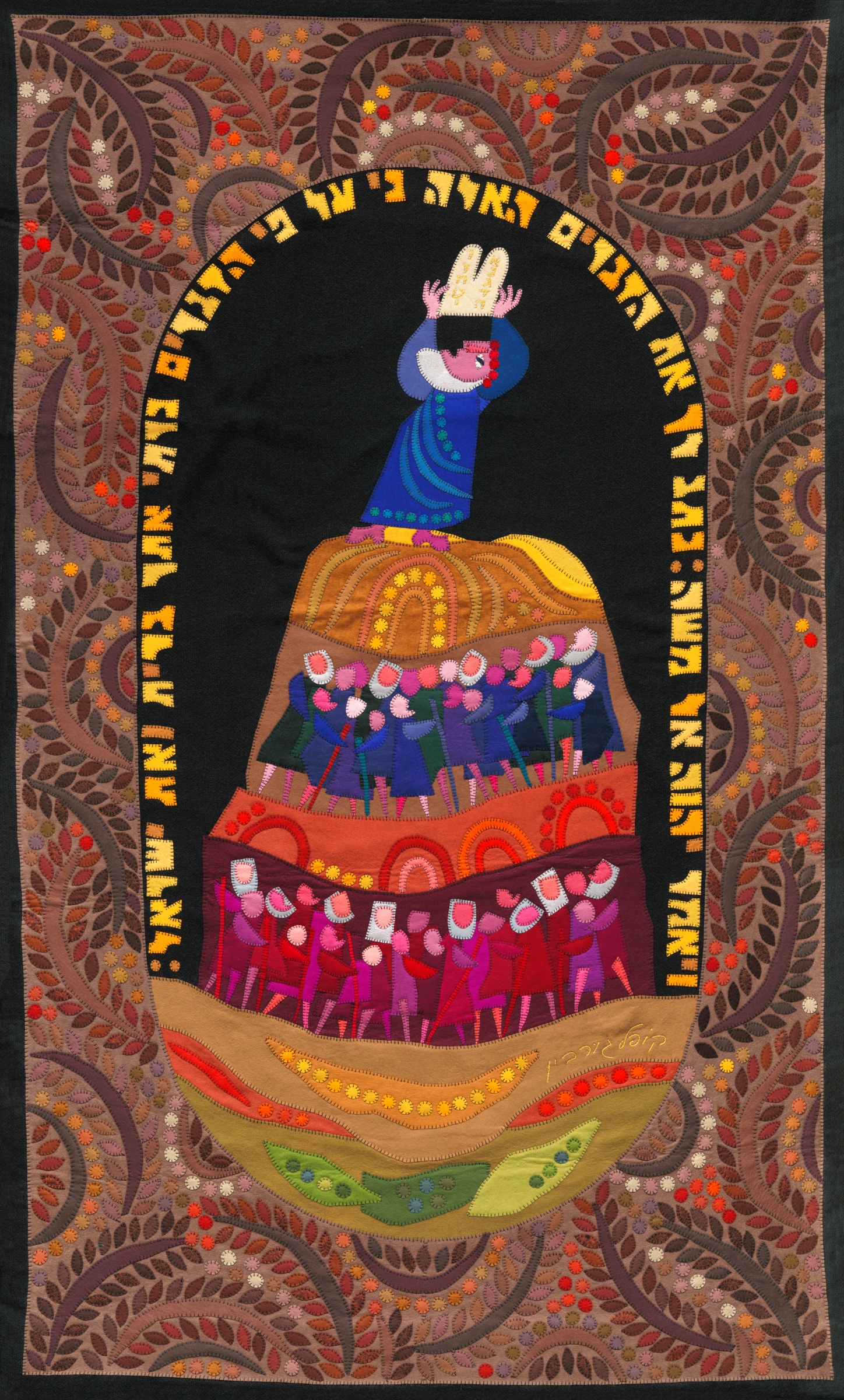 The Revelation on Mount Sinai by Kopel Gurwin, a hand-cut felt appliqué depicting Moses raising the Tablets of the Covenant above a gathering of Israelites arranged in colorful tiers. The central scene is enclosed by an arch of Hebrew lettering and an elaborate ornamental border of branching vines, leaves, and flowers in earthy brown, orange, and pink tones.