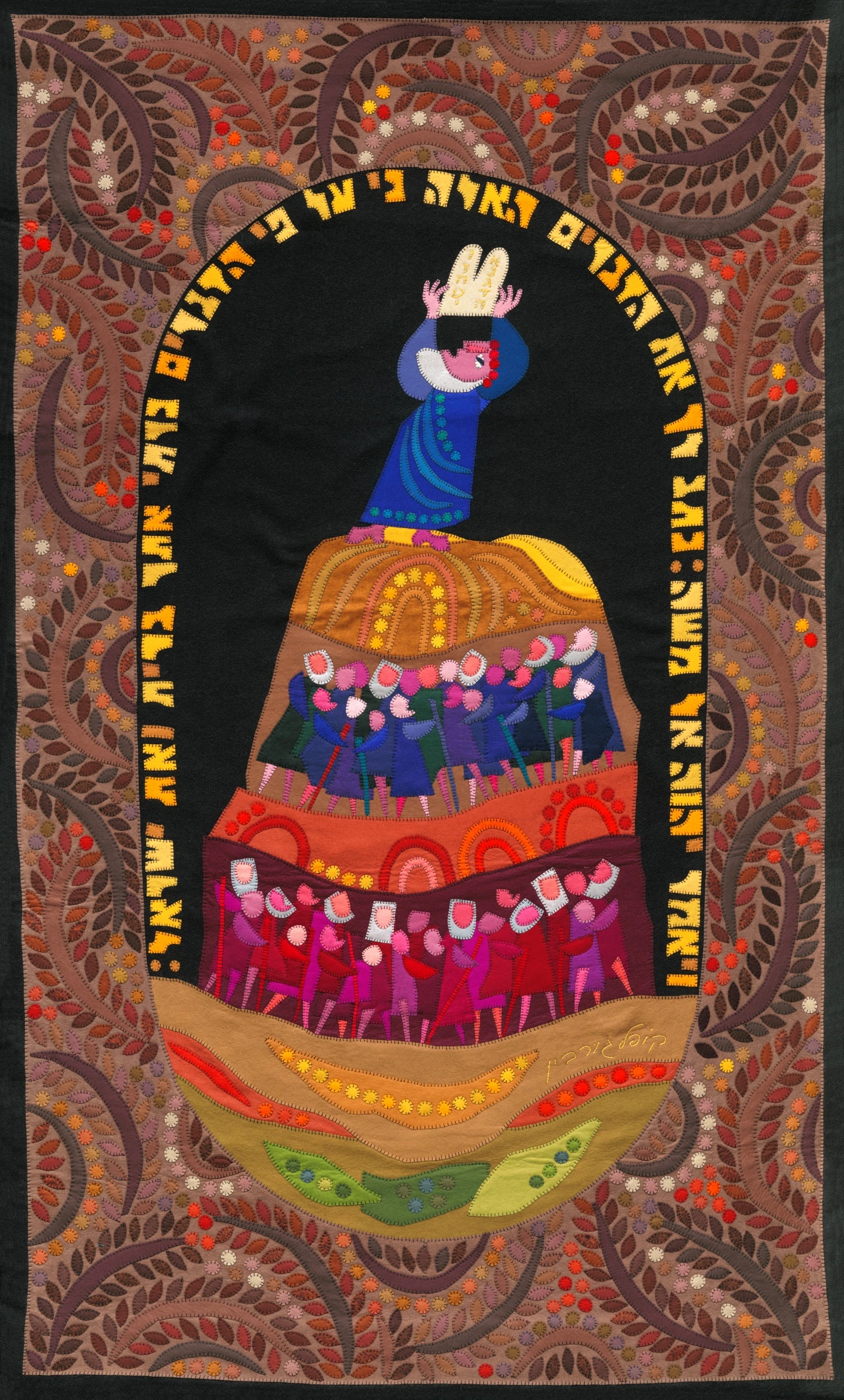 The Revelation on Mount Sinai by Kopel Gurwin, a hand-cut felt appliqué depicting Moses raising the Tablets of the Covenant above a gathering of Israelites arranged in colorful tiers. The central scene is enclosed by an arch of Hebrew lettering and an elaborate ornamental border of branching vines, leaves, and flowers in earthy brown, orange, and pink tones.