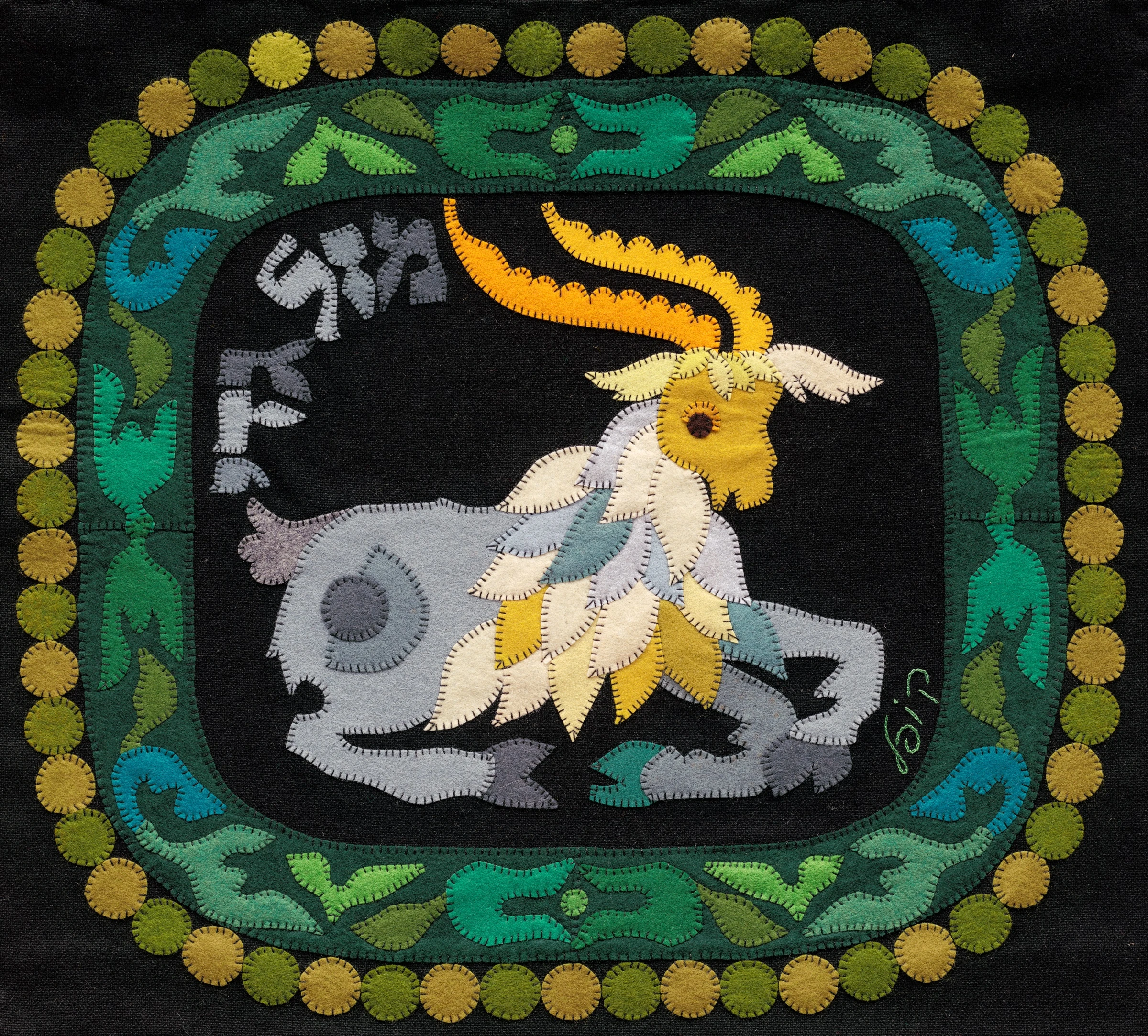 Capricorn by Kopel Gurwin, a hand-cut felt appliqué depicting a horned goat in pale grey, cream, and yellow against a black ground, framed by green leaf-like ornamentation and a border of circular forms. The Hebrew inscription for Mazal Gdi appears in the upper left.